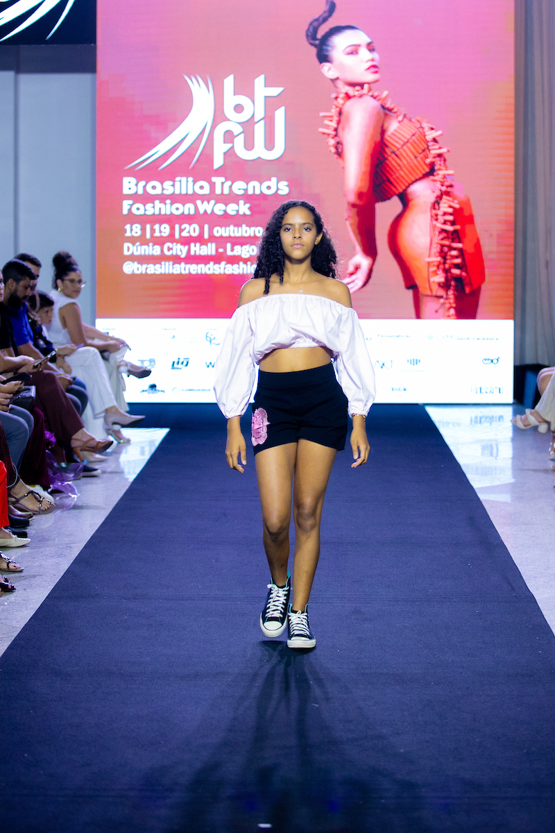 A imagem mostra uma modelo mirim desfilando em uma passarela. Em frente à um telão do evento, a jovem modelo caminha com um top branco e bermuda preta