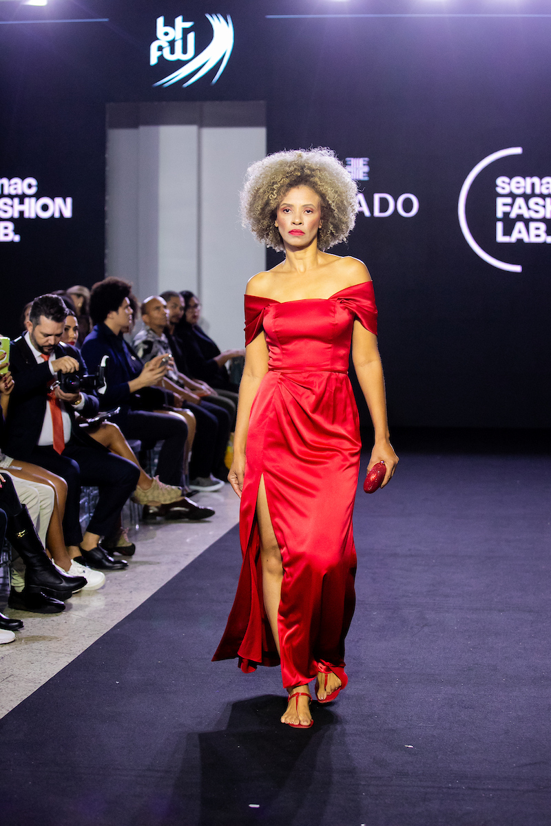 A imagem mostra uma modelo desfilando. Com um vestido vermelho, a mulher negra caminha sob a passarela olhando para a frente