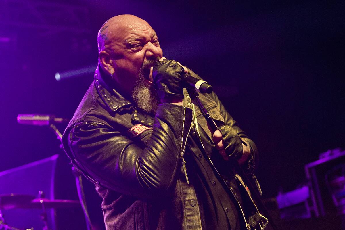 Imagem colorida de homem careca cantando em microfone. Ele usa jaqueta de couro - Metrópoles