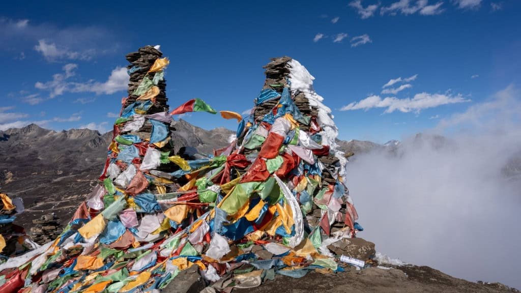 Duas pilhas de pedras estão cuidadosamente empilhadas em um terreno rochoso, cobertas por bandeiras coloridas de oração tibetanas que esvoaçam com o vento. Ao fundo, imponentes montanhas parcialmente encobertas por névoa compõem uma paisagem fria e elevada.