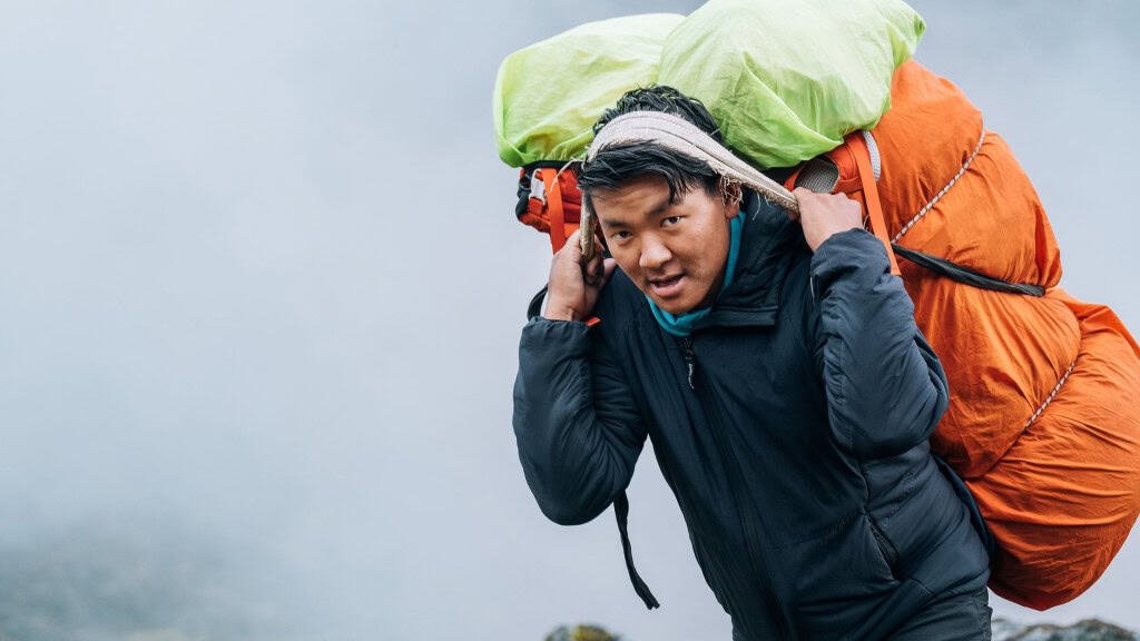 Uma pessoa caminha em direção à neblina, carregando duas mochilas grandes — uma verde e outra laranja. O cenário é montanhoso, envolto por nuvens baixas que sugerem esforço e resiliência. Com uma jaqueta escura, ela avança com determinação, como quem enfrenta uma jornada exigente.