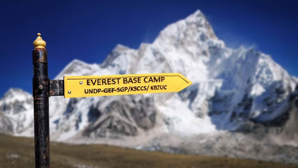 Uma placa amarela aponta para o Acampamento Base do Everest, com letras pretas e fundo de montanhas nevadas. Atrás dela, o horizonte exibe picos imponentes do Himalaia sob um céu azul claro. O contraste entre o sinal e a natureza transmite a grandiosidade e o desafio da jornada.