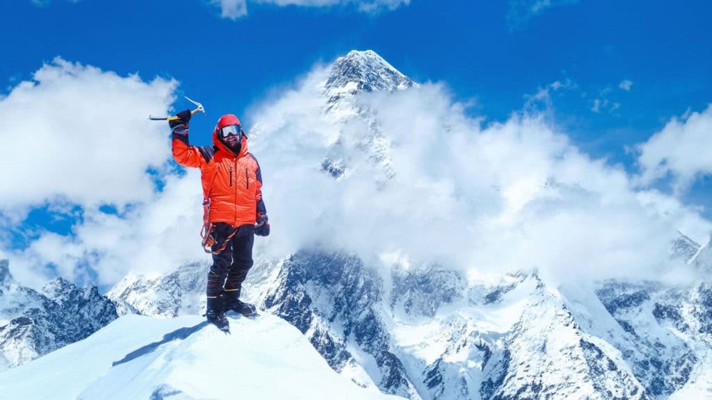 No topo de uma montanha coberta de neve, uma pessoa ergue seu machado de gelo como símbolo de conquista. O céu azul e as montanhas ao fundo criam um cenário grandioso e sereno. A jaqueta laranja vibrante destaca sua presença num momento de superação e liberdade.
