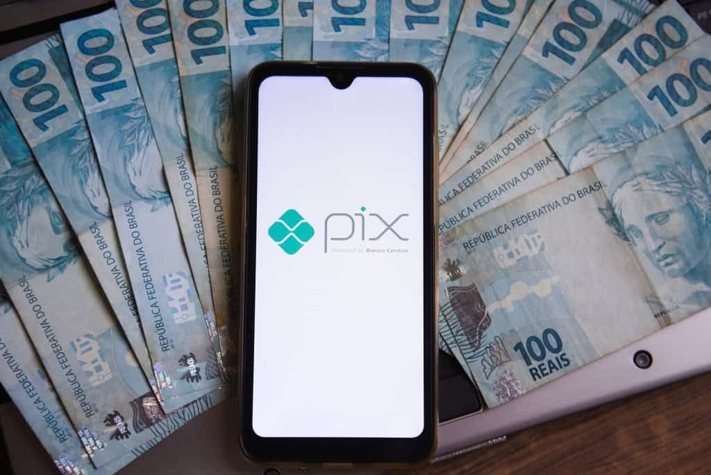 Notas de R$ 100 espalhadas; por cima, um smartphone com o logotipo do Pix
