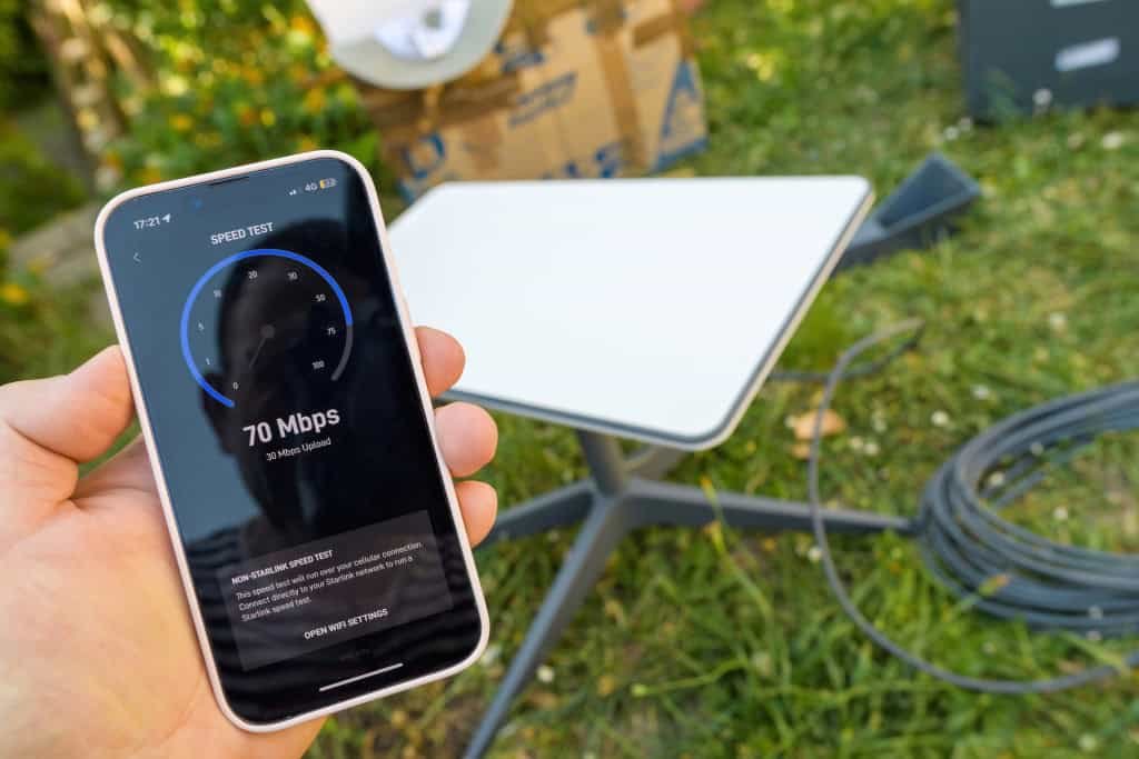 Ao fundo, antena da Starlink em um gramado; acima, pessoa segura um smartphone medindo a velocidade da internet