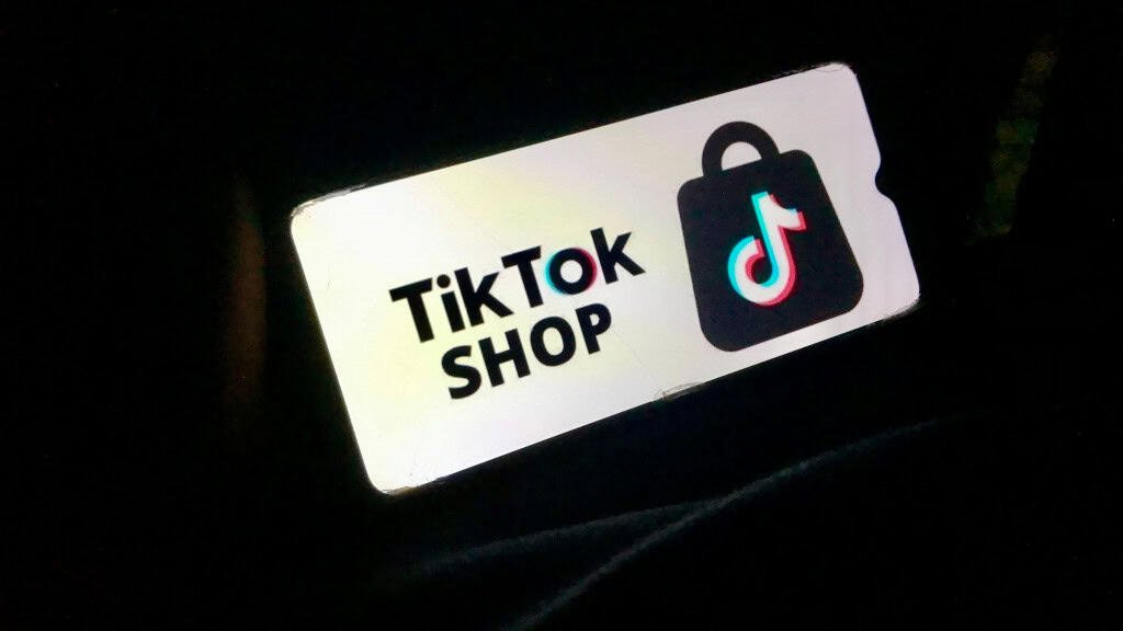 A imagem mostra um celular exibindo o logo e o texto "TikTok Shop" em uma tela branca, destacando-se de um fundo escuro. Basicamente, é uma representação visual do TikTok Shop, a funcionalidade de comércio eletrônico do TikTok. O TikTok Shop foi lançado no Brasil em maio de 2025 e permite que os usuários comprem produtos diretamente no aplicativo, através de vídeos, lives e perfis de lojas, com influenciadores sendo comissionados pelas vendas.