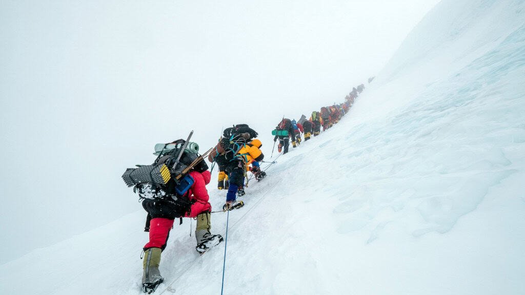 Uma longa fila de alpinistas sobe uma encosta íngreme e coberta de neve, conectados por cordas para segurança. Cada pessoa carrega mochilas pesadas e equipamentos de escalada, avançando com esforço em meio ao gelo e altitude extrema. A imagem transmite trabalho em equipe, superação e os desafios intensos do montanhismo em grandes altitudes.