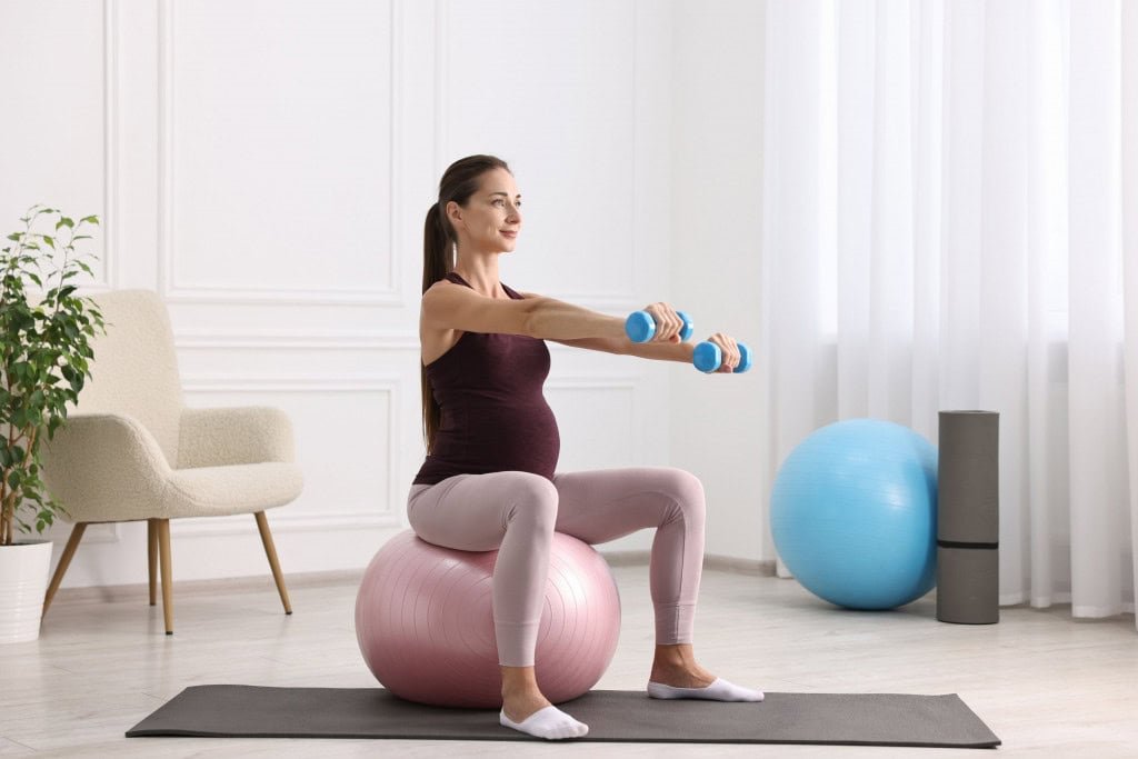 Grávida praticando atividade física, sentada em bola de pilates e com halteres nas mãos