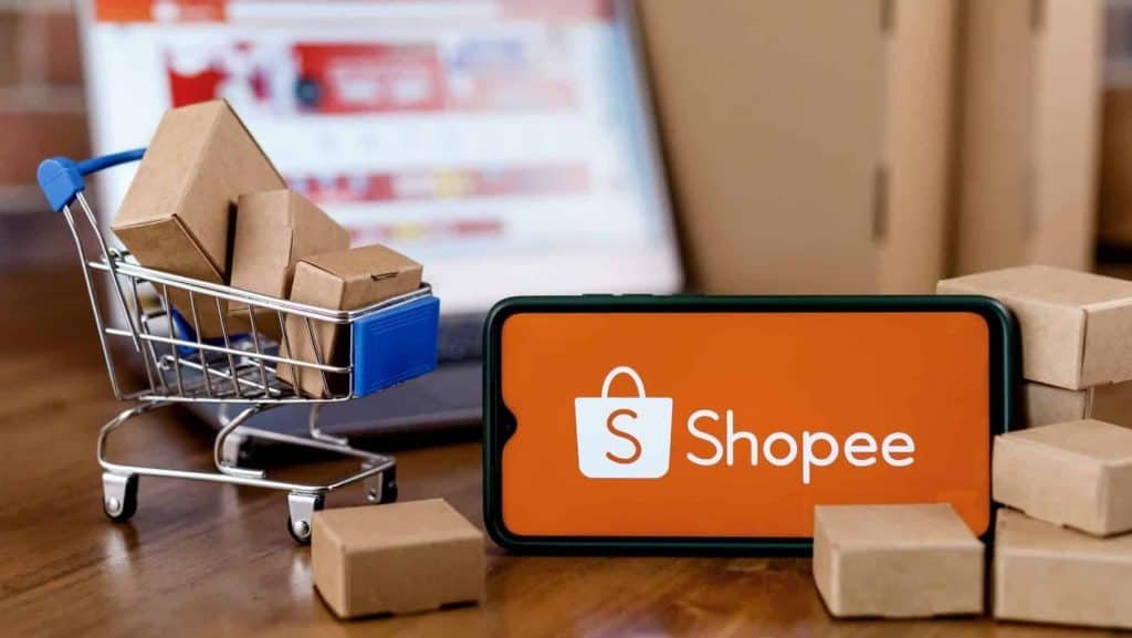 Logo da Shopee em um smartphone ao lado de um mini carrinho de compras com caixinhas dentro e ao lado
