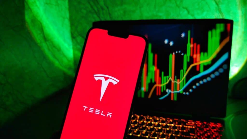 À frente, logo da Tesla em um smartphone; ao fundo, um notebook com gráficos